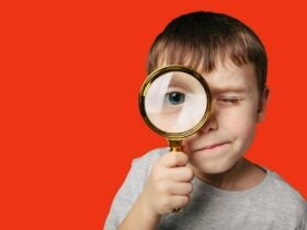 A boy standing front of a red background looks through a magnifying glass.