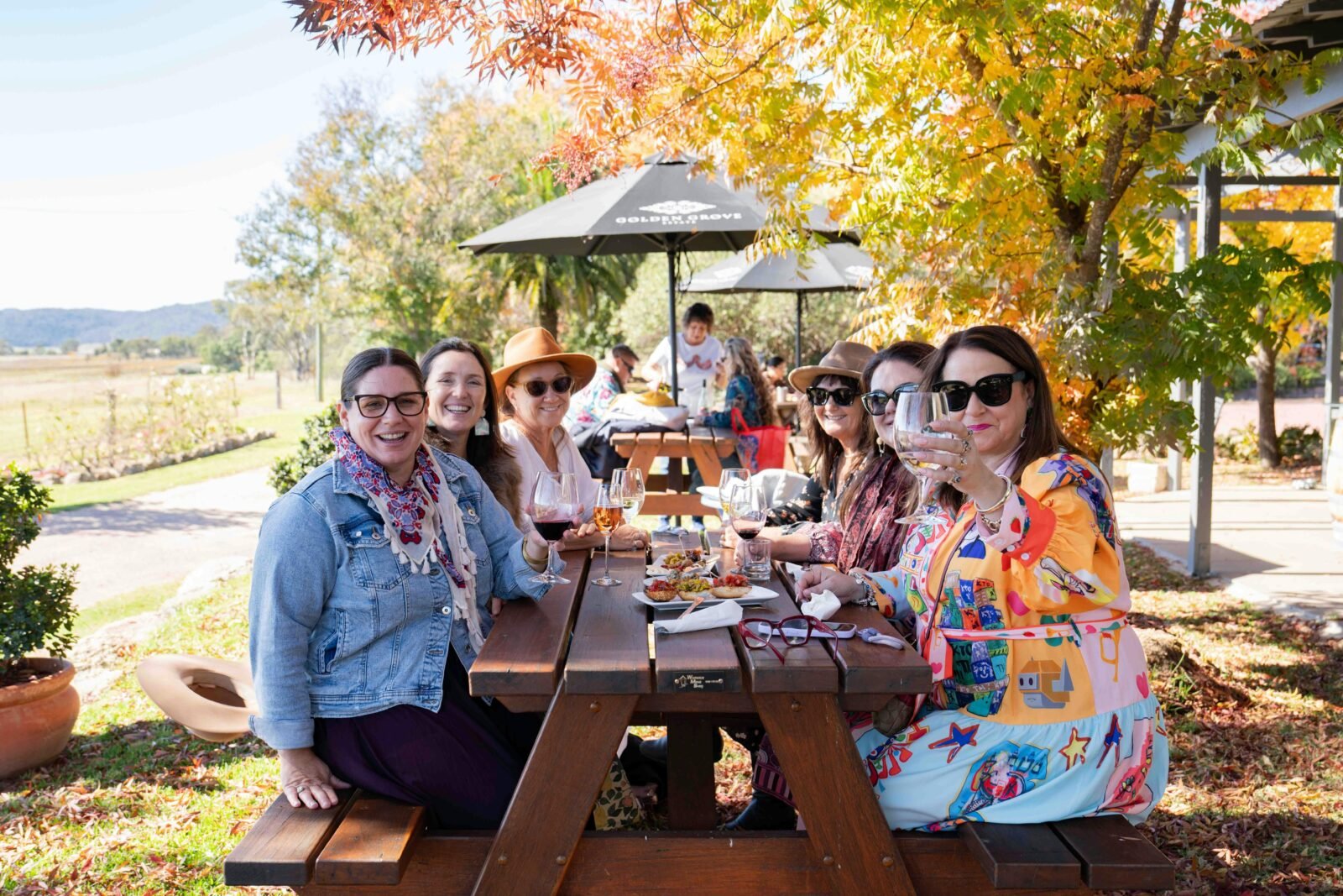 A group of women sitting at wooden table under an orange leafed tree