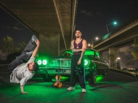 Lowrider car under overpass with neon lights, dancers, and violin on ground.