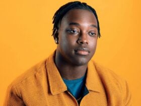 Black man with short twists & mustard jacket over teal shirt, posing against solid orange background