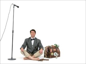 A man in a tuxedo shirt and jacket with shorts sitting on the ground between a mic and a suitcase