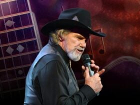 A man in a black vest and cowboy hat, singing into a microphone