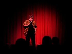 Me with short hair in a yellow flannel shirt standing in front of a red curtain talking into a mic