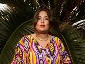 White person in colourful geometric-patterned dress and red lipstick, stands in front of palm leaves