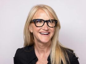 Mel Robbins pictured close up wearing black rim glasses and a black top and long blonde hair smiling