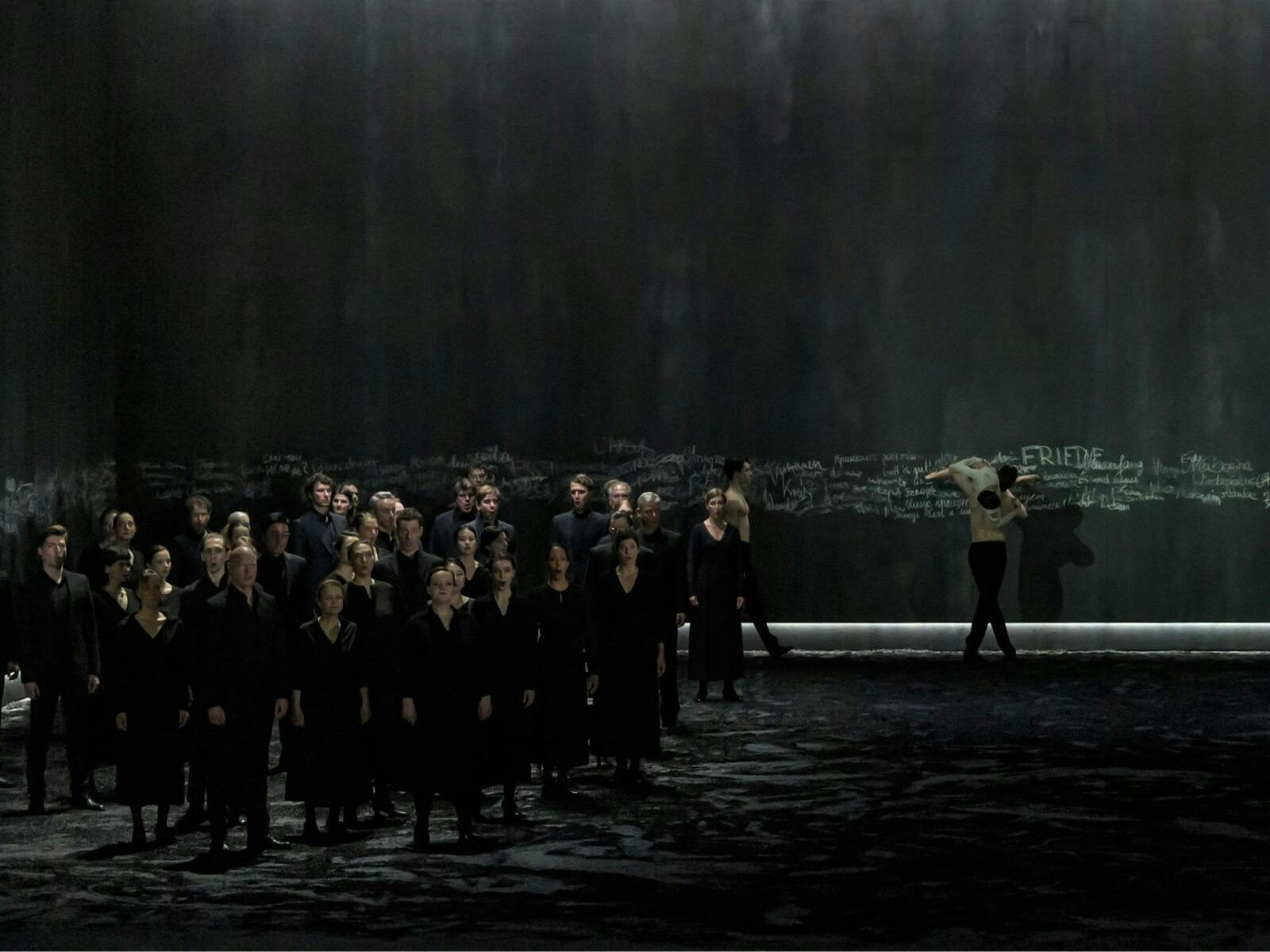 Large choir on stage with dancer couple on the right. Dark grey wall at the back of the image.