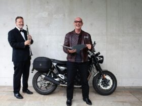 A smiling man in a leather jacket holds a folder in front of a motorcycle.