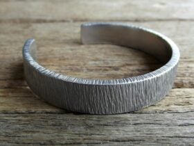 Silver-smithed bracelet with textured pattern displayed on wooden bench.