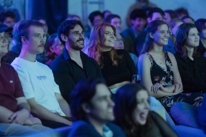 A group of people are seated in an audience, smiling and engaged.