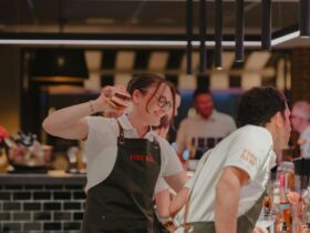 Smiling bartender in Fire Baby apron shaking a cocktail behind a busy bar, warm lighting and lively