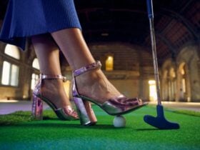 Close-up of a woman wearing pink heels, standing on indoor green turf beside a golf club and ball.