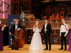 Six classical musicians on stage: violinist, double bassist, soprano, pianist, and flautist.