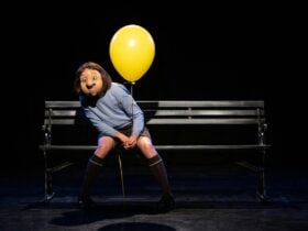 An actor with a face mask sitting on a park bench holding a yellow balloon