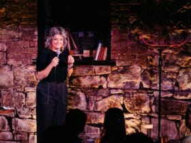 Comedian Chris Ryan on stage at a comedy club, with a heritage brick wall behind her.