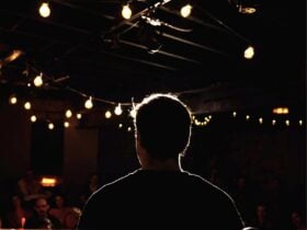 Photo taken from behind of a comedian on stage, their figure silhouetted in front of an audience.