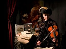 A man dressed in rich velvet cloak and feathered hat playing a violin in front of cluttered table