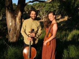 Two people standing outdoors holding a cello and a violin in warm sunlight.