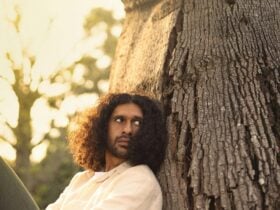 A man is sitting and resting against a big tree trunk. He is looking over his shoulders and waiting.