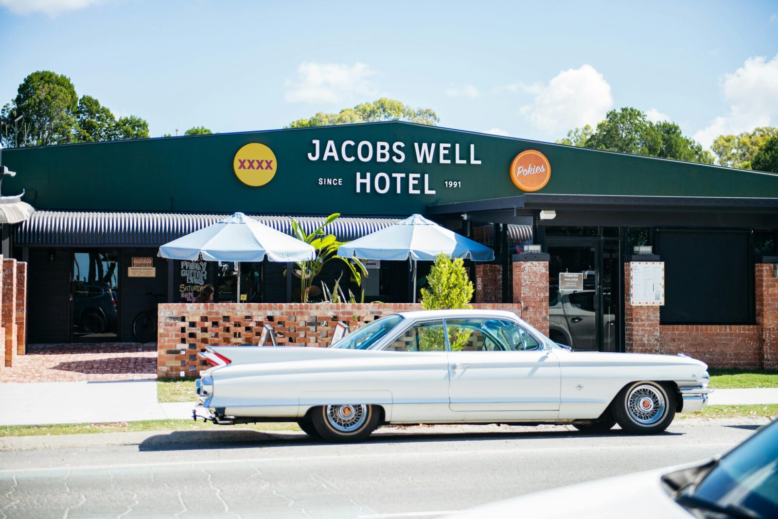 A vintage white Cadillac parked in front of the dark green Jacobs Well Hotel building on a sunny day