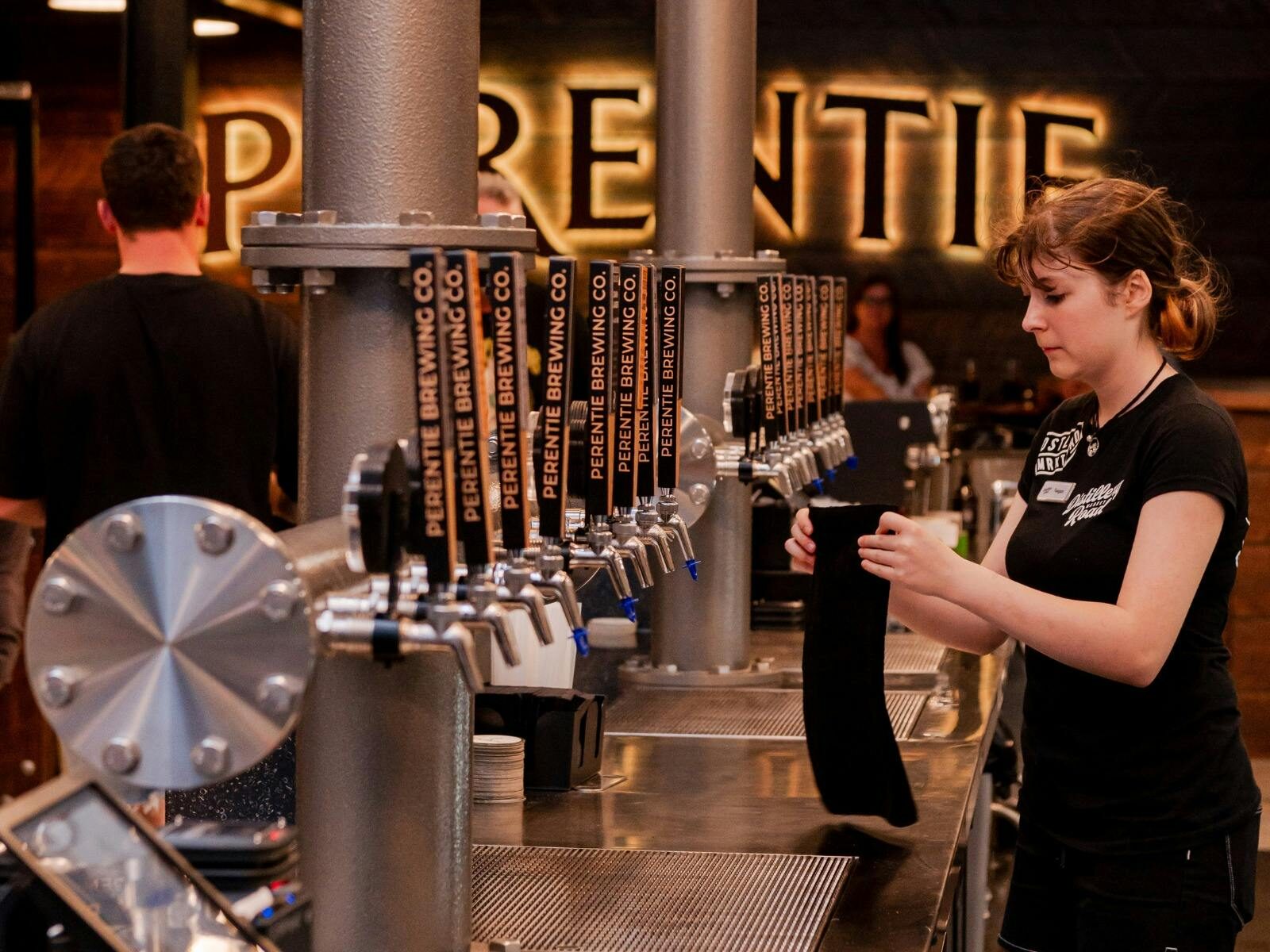 Young female stands behind bar where a row of beer taps can be seen in front of a Perentie sign