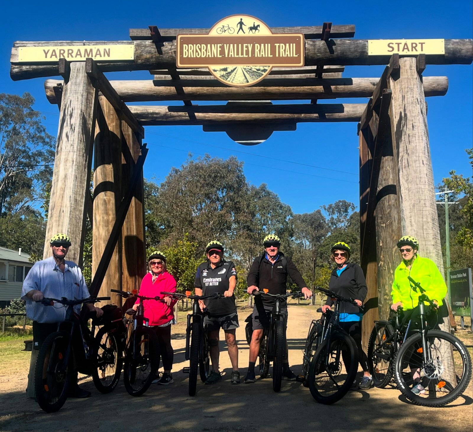 A group of 6 riders stand in front of a wooden structure, they are holding bikes and wearing helmets