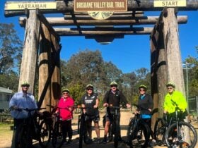 A group of 6 riders stand in front of a wooden structure, they are holding bikes and wearing helmets
