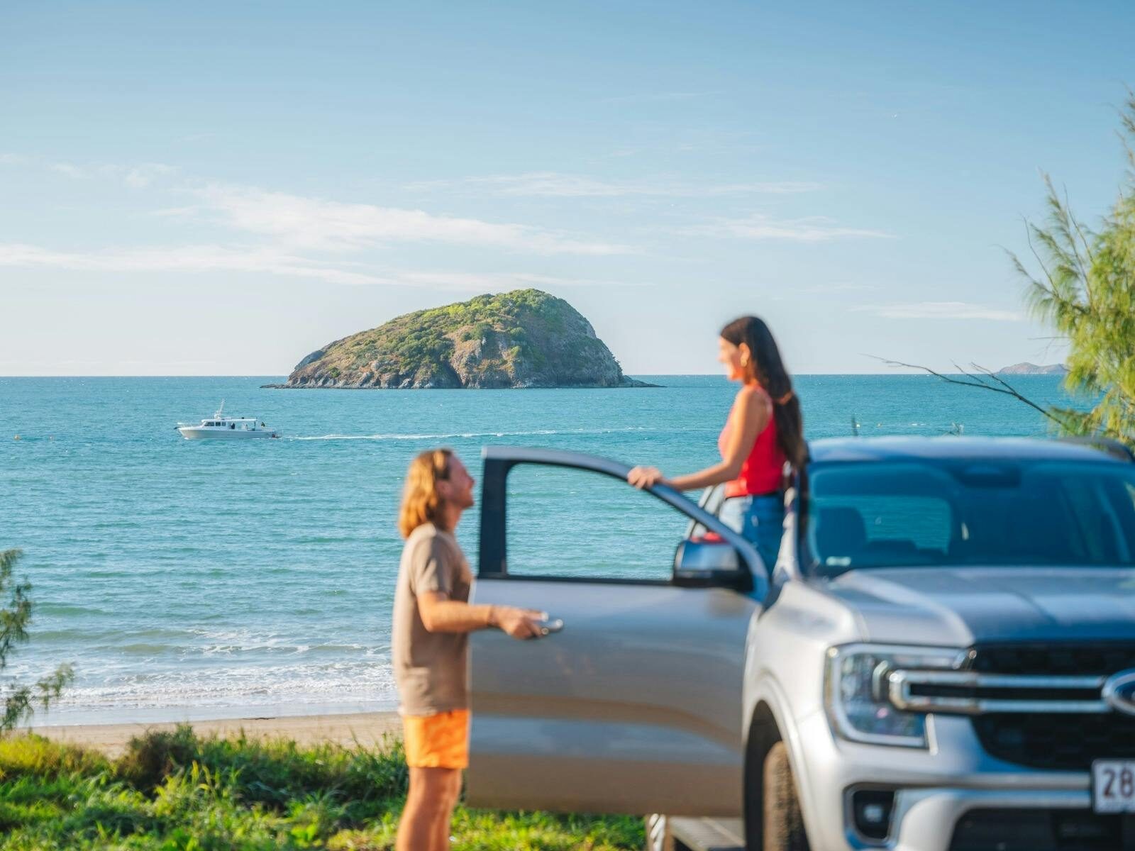Two tourists pause to look at a boat and a distant mountain rising from the sea