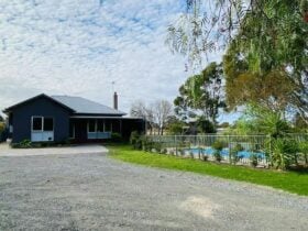 Modern dark-grey house with large windows, gravel driveway, and fenced pool surrounded by lush green