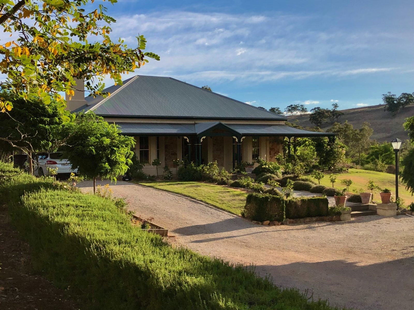 A country homestead, with gardens and trees surrounding it. In the distance are rolling hills.