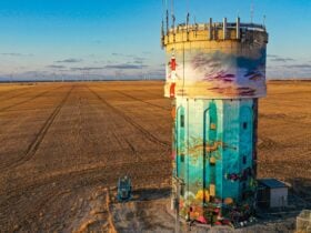 Tall water tower with ocean mural, set in flat farmland with wind turbines on horizon.