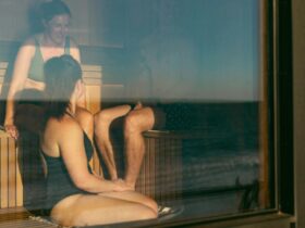 Two female and one male bather enjoy the sauna, smiling as the sun shines through the sauna window.