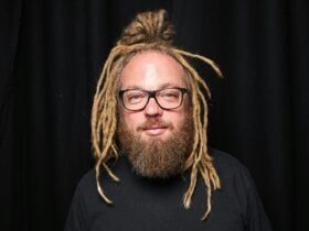 Fabien Clark - Comedian with dreadlocks, beard and glasses against a black background