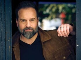 A bearded man in a brown suede jacket stands in a dark doorway looking directly at the camera.