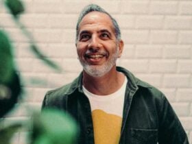 Portrait of chef Yotam Ottolenghi looking up and smiling, with beige brick wall in the background