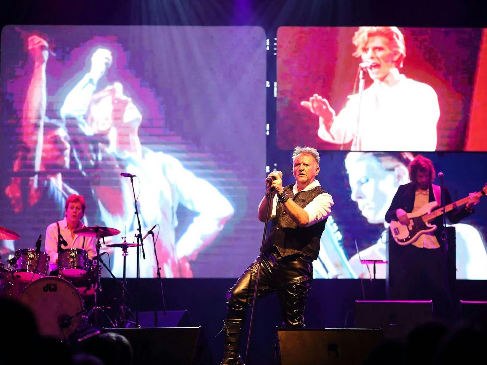 Male singer and two musicians in the background performing in front of screen images of David Bowie