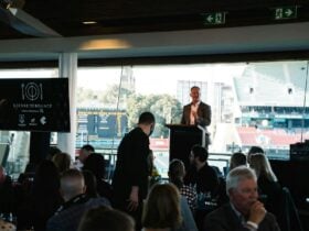 Man standing at microphone in front of a room full of people at a large oval