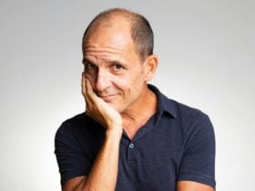 Comedian Carl Barron smiles, wearing a dark blue polo shirt, resting his head on his hand