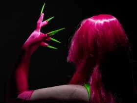 An AFAB figure shot from behind poses with a hot pink glove with long green nails next to their head