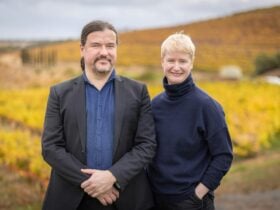 Two people stand and smile in front of grapevines in McLaren Vale