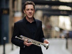 Trumpeter David Elton standing with his trumpet in a blurred street.
