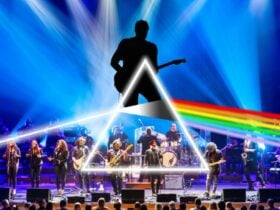 A live band performs on stage under blue lighting with a prism graphic projecting a rainbow