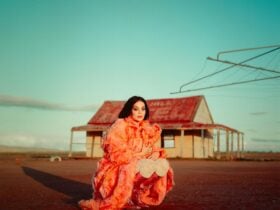 A lady with long back hair sitting in a rural setting wearing a long peach dress and high red boots