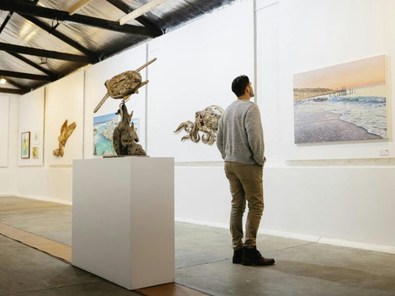 A man in a grey jumper and beige pants in an art gallery looking at a picture of a beach