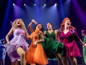 Four women in colourful, sparkly dresses dance enthusiastically on stage under bright spotlights