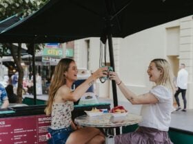 Two girls enjoy a smashburger and fries at a table while they cheers their beer together.