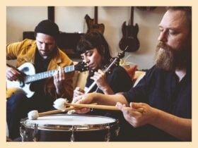 Three musicians play guitar, strings, and drum together in a cozy indoor setting.