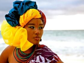 Grace Barbé, wearing a yellow and blue headdress, standing in front of the ocean.