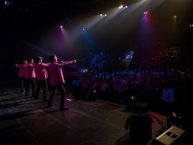 Onstage view of four pink-jacketed singers performing to a large seated theater crowd applauds loud