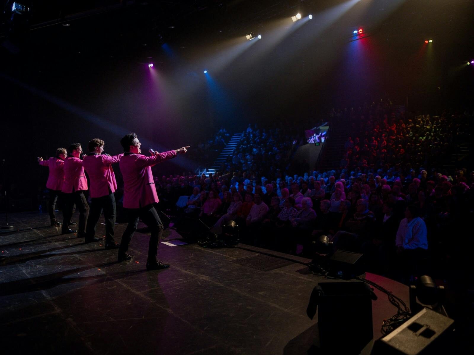 Onstage view of four pink-jacketed singers performing to a large seated theater crowd applauds loud