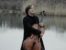 Ivan Karizna holding his cello and staring into middle distance in front of a lake.